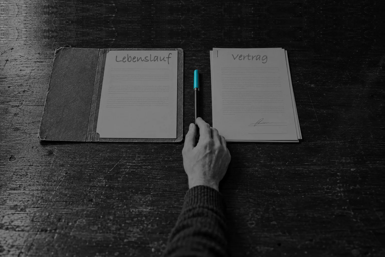 Auf einem dunklen Schreibtisch liegen nebeneinander eine offene Bewerbungsmappe mit einem Blatt, auf dem von Hand „Lebenslauf" steht, und ein gehefteter Vertrag mit der Überschrift „Vertrag", auf dem eine Unterschrift steht und die zweite Unterschriftszeile leer ist. Dazwischen liegt ein geschlossener Stift mit cyanfarbener Kappe, den eine Hand mit den Fingerspitzen berührt, ohne ihn aufzunehmen.