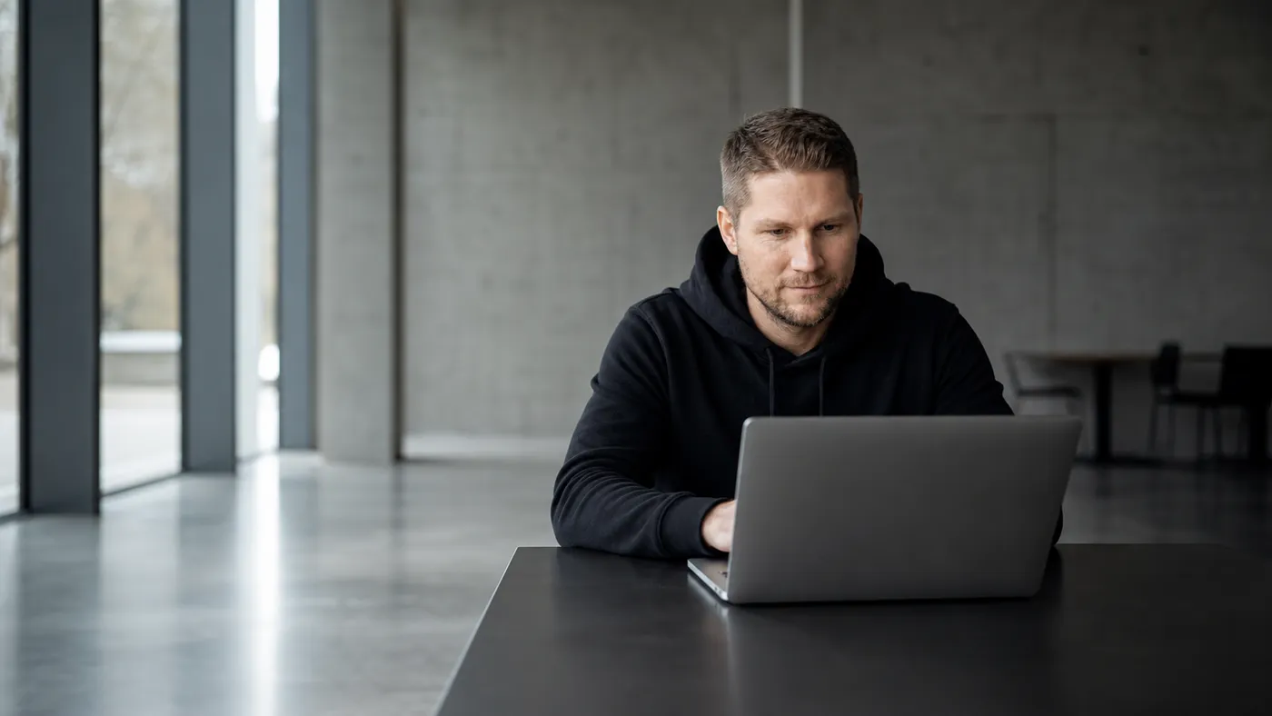 Ein Mann in schwarzem Hoodie sitzt allein an einem dunklen Tisch vor einem aufgeklappten Laptop und liest konzentriert vom Bildschirm. Der Raum dahinter ist leer, im Hintergrund eine Betonwand und eine Fensterfront.